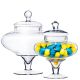 Two clear glass apothecary jars with pedestal bases and lids, one empty and the other filled with blue and yellow gumballs.